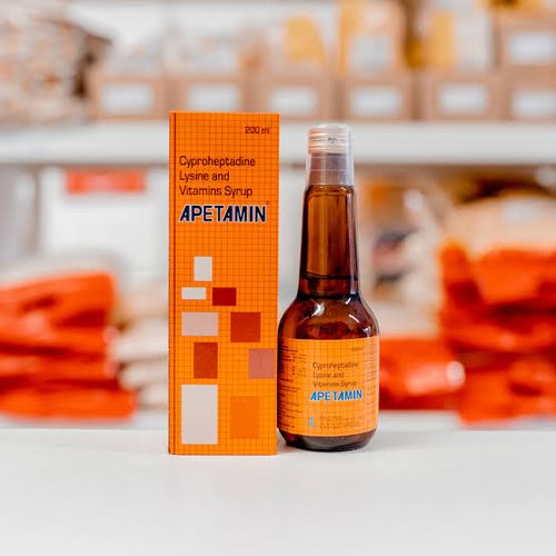 Orange box and brown bottle of Apetamin syrup on a white surface with blurred background.