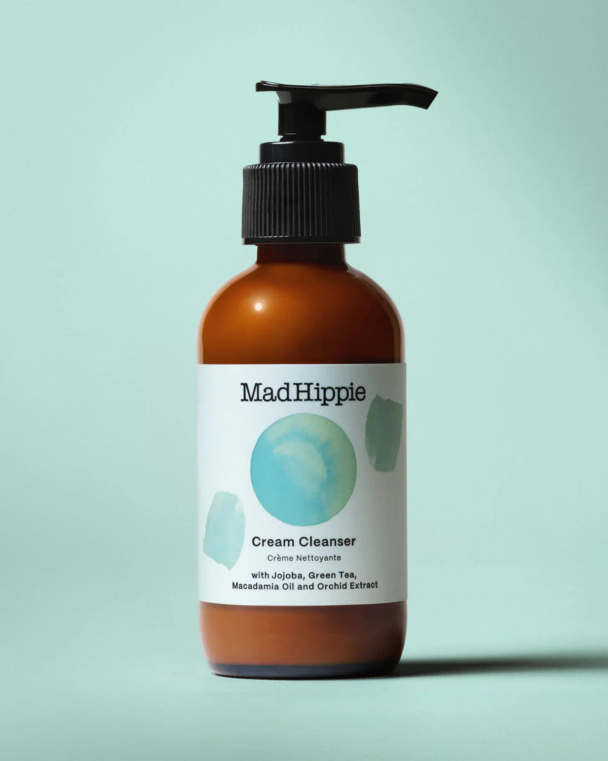 Amber bottle with black pump, labeled "MadHippie Cream Cleanser" on a light teal background.